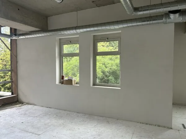 Binnenruimte in verbouwing met twee ramen, zicht op groen buiten en zichtbare ventilatiebuizen aan het plafond.